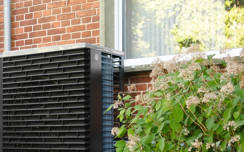 Zwarte warmtepomp-buitenunit tegen de baksteengevel van een pand